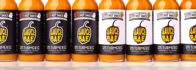 Lineup of Sauce Bae hot sauces featuring alternating bottles of Hotter Habanero with black labels and Skinny Habanero with white labels. Each 5 oz bottle displays the Sauce Bae logo and “Made with Turmeric” tagline. The hot sauces are arranged in a clean row against a white background with subtle reflections below, showcasing the visual contrast between the two flavors.